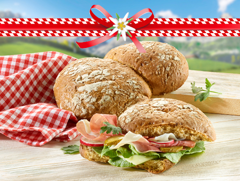 Rustikale Brötchen mit Schinken, Salat und Käse vor Alpenkulisse mit Deko-Schleife.