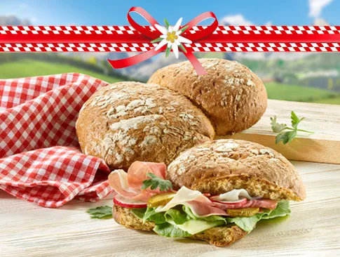 Rustikale Brötchen mit Schinken, Salat und Käse vor Alpenkulisse mit Deko-Schleife.